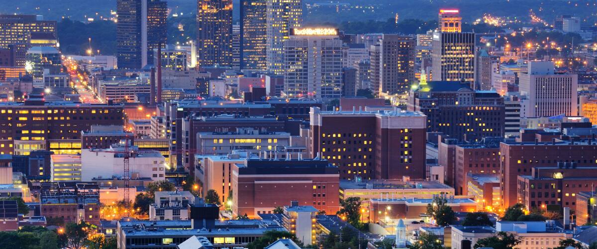 A cityscape at dusk with illuminated buildings and a mix of modern and historic architecture.