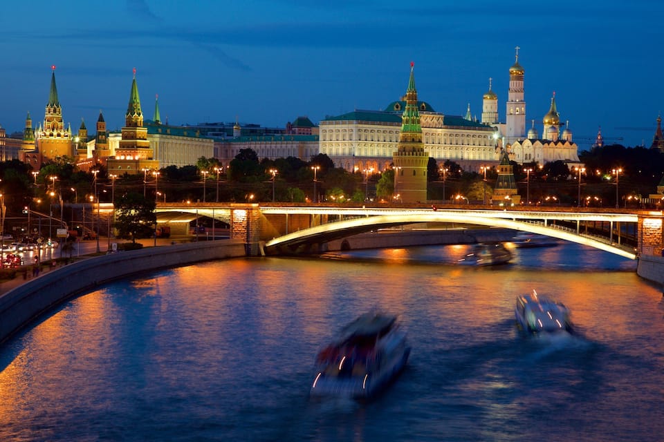 A nighttime view of a river with a bridge, a historic building with domes, and a tower in the background.