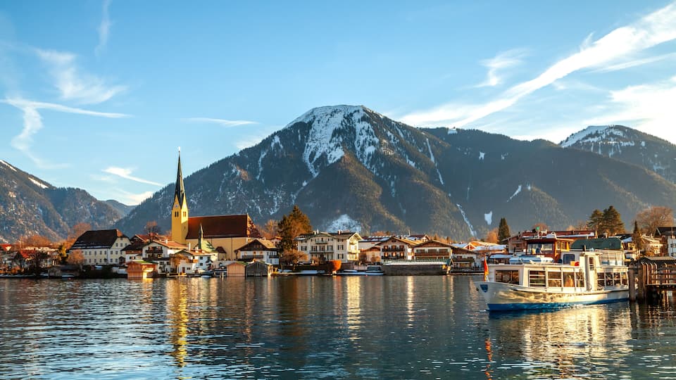 A lakeside town with a church, boats, and snow-capped mountains in the background.