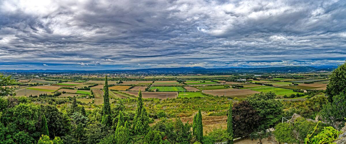 A panoramic view of a rural landscape with fields, trees, and distant mountains under a cloudy sky.