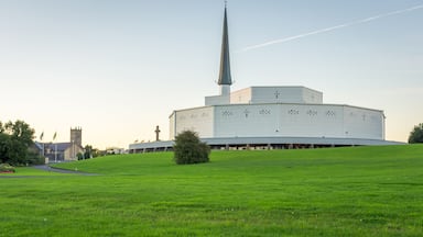 Un edificio moderno con una aguja alta, rodeado de un campo verde y una iglesia al fondo.