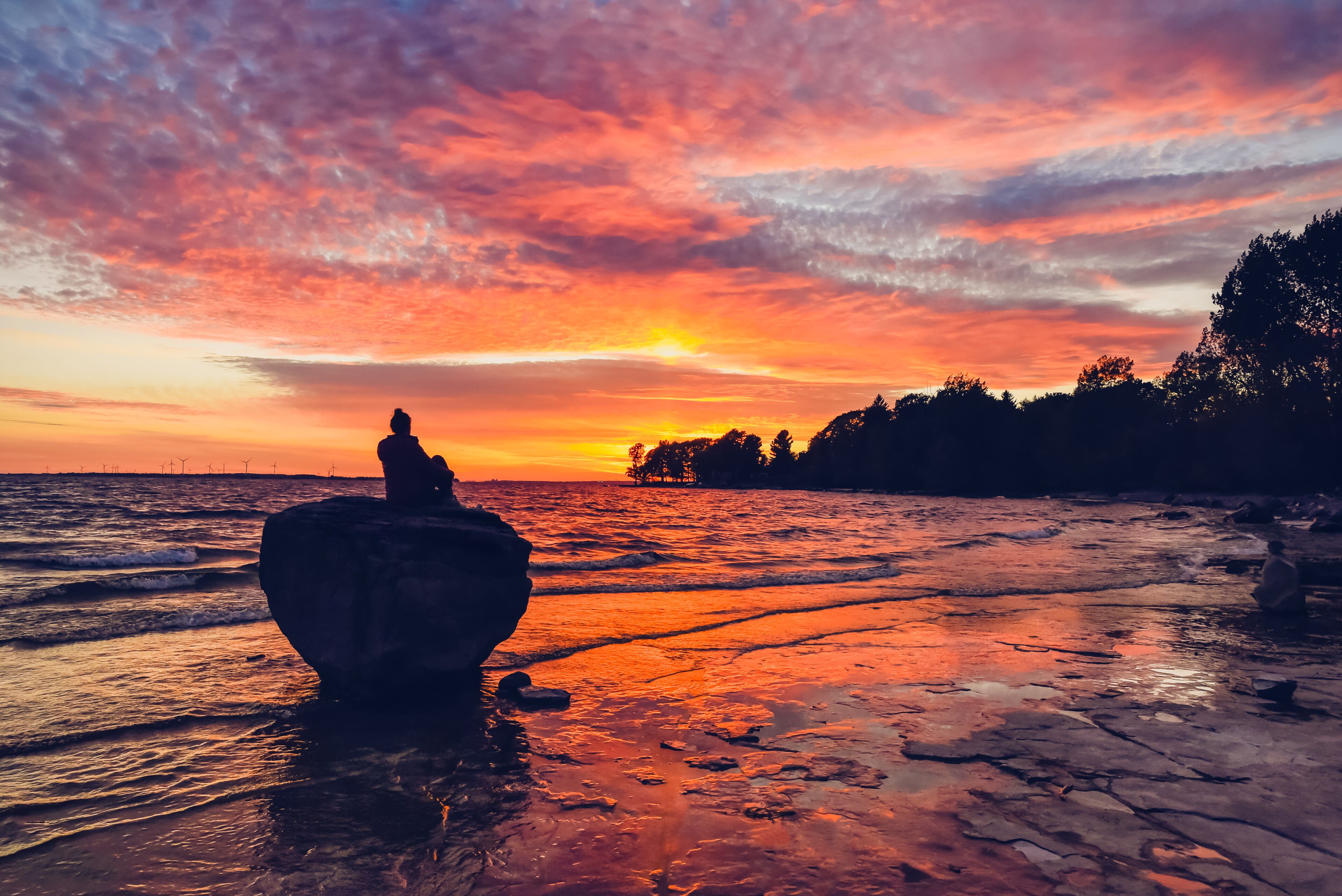 A sunset over a rocky beach with a silhouette of a person on a rock and wind turbines in the distance.