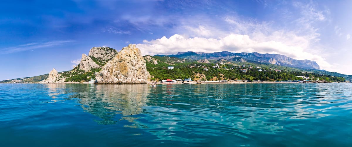 A coastal landscape with a prominent rocky outcrop, clear turquoise waters, and a backdrop of mountains under a partly cloudy sky.