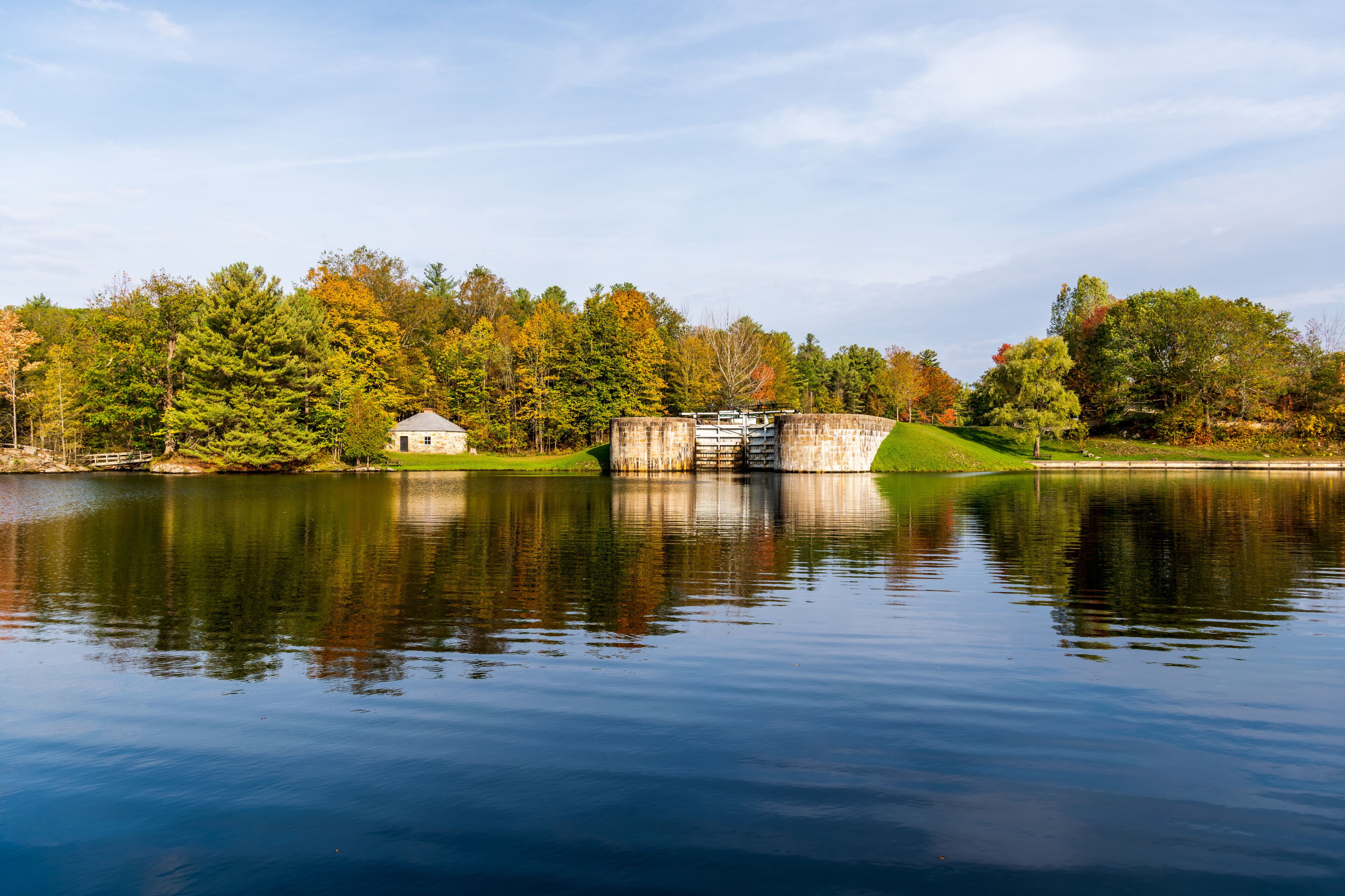 A calm lake with a stone dam, a small house, and autumn-colored trees in the background.