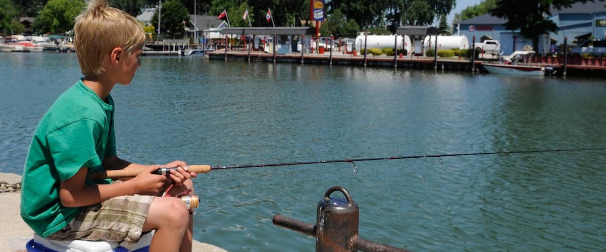 A boy fishing by a dock with a boat in the background.