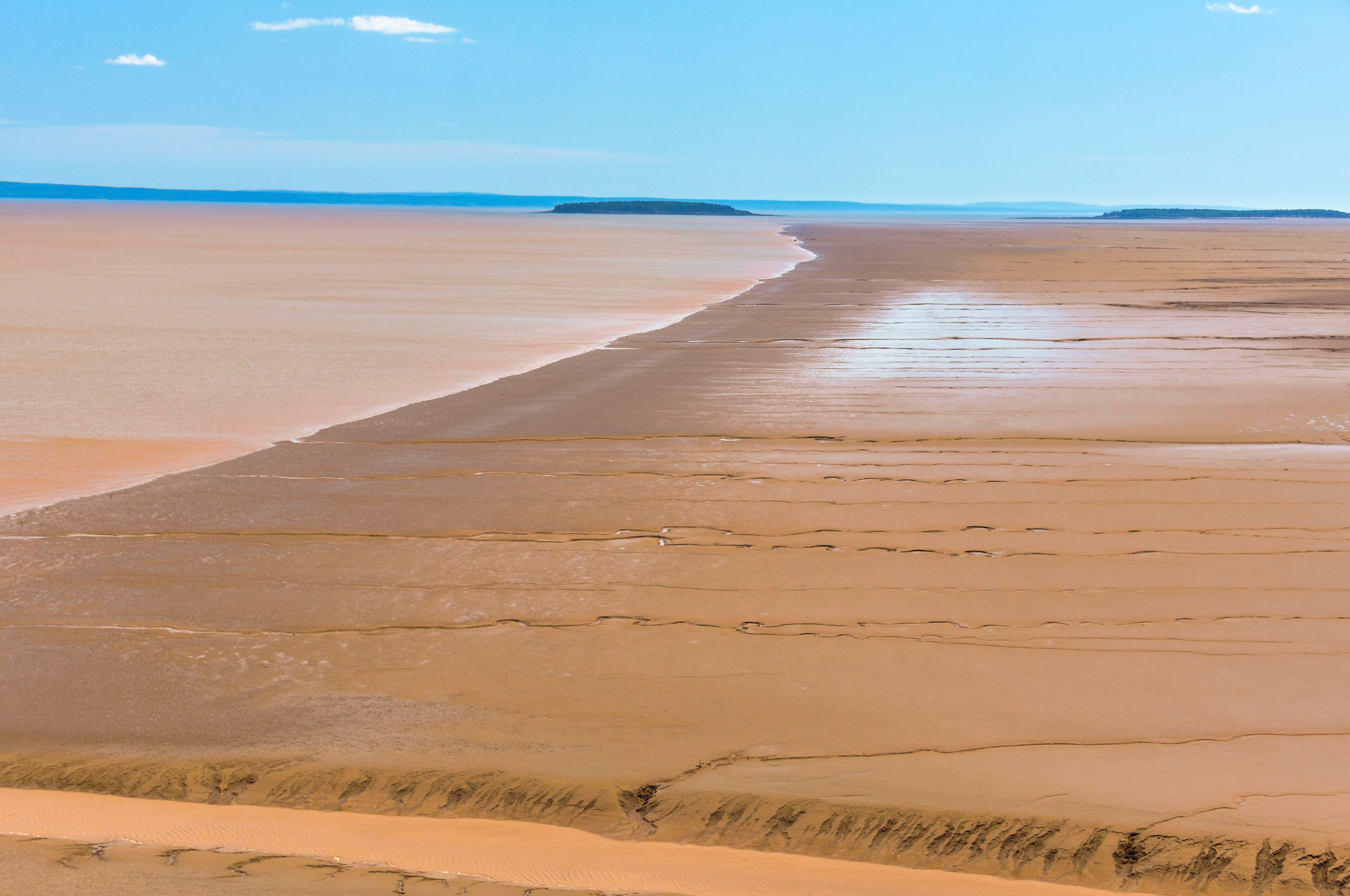 A vast, flat sandy beach with gentle waves lapping at the shore, a clear sky, and distant landforms.