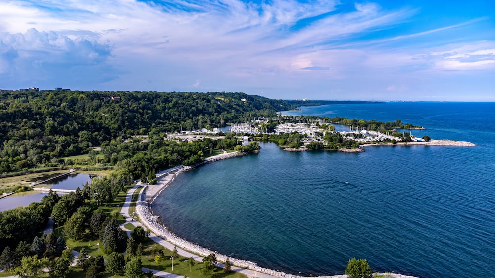 Une ville côtière dotée d’un port de plaisance, entourée de collines boisées et surplombée d’un ciel bleu azur.
