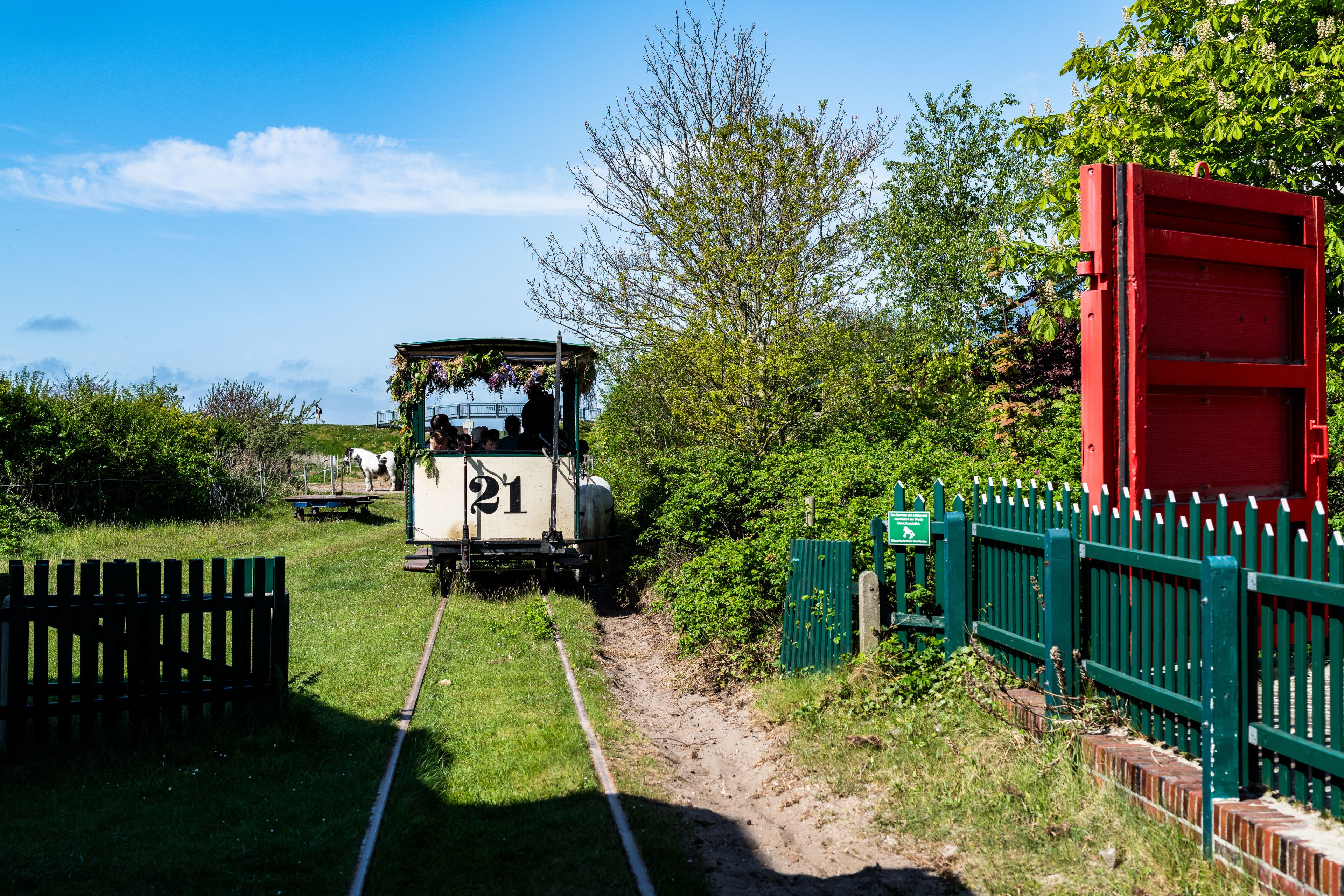 A narrow railway track with a vintage train labeled '21' approaching a green fence and a red structure.