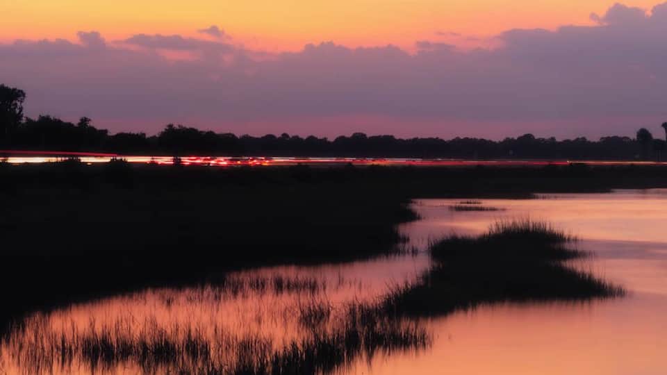 A sunset over a calm water body with reflections of the sky, surrounded by grassy areas and distant trees.