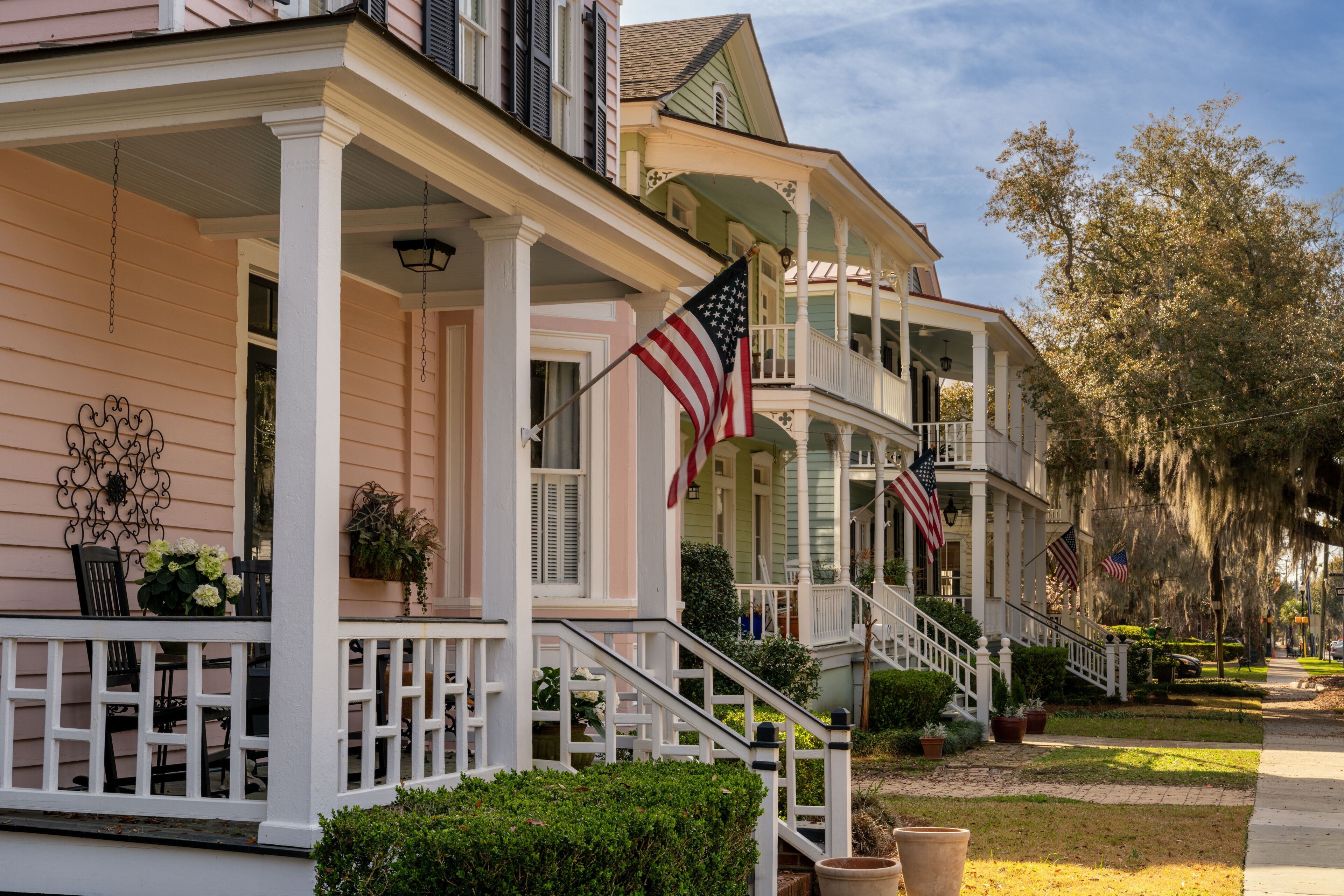 A row of pastel-colored houses with American flags, white railings, and a well-maintained lawn.