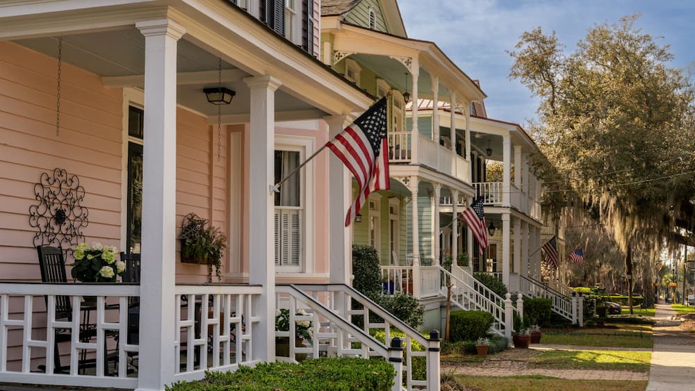 A row of pastel-colored houses with American flags, white railings, and a well-maintained lawn.