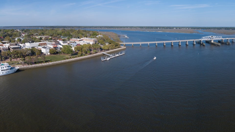 A river with a bridge, a white boat docked on the riverbank, and a town with buildings and trees.