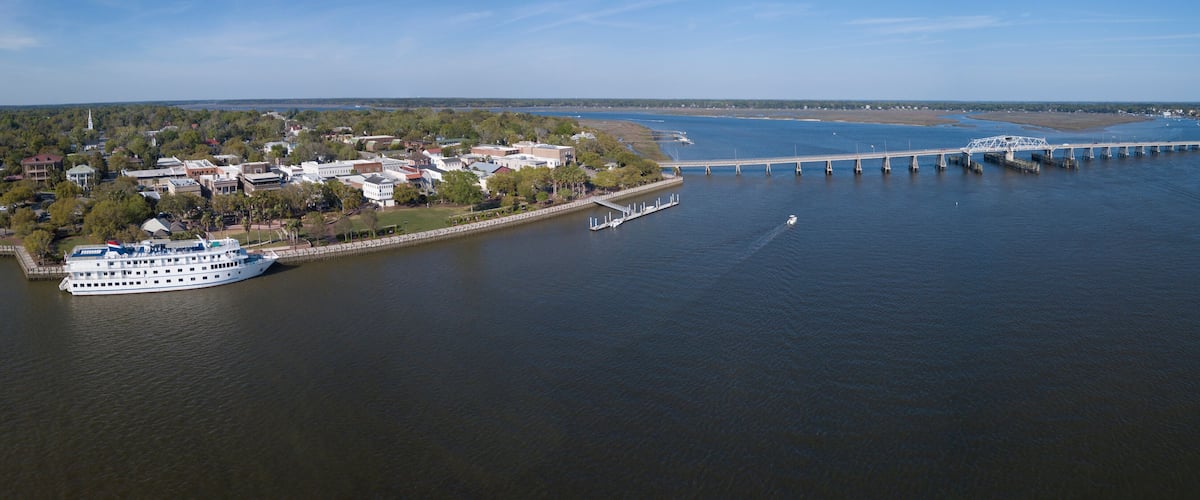 A river with a bridge, a white boat docked on the riverbank, and a town with buildings and trees.