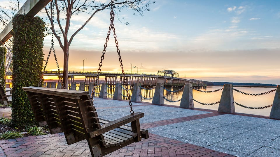 A wooden swing bench overlooking a waterfront with a bridge in the background.