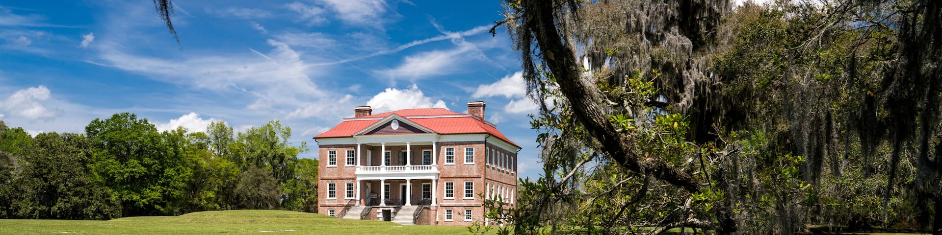 A large brick building with a red roof, a pond in the foreground, and trees with Spanish moss.