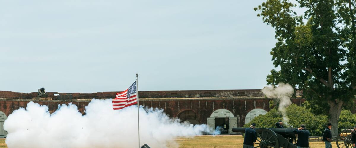 A cannon firing with smoke, an American flag, and a historic fort in the background.