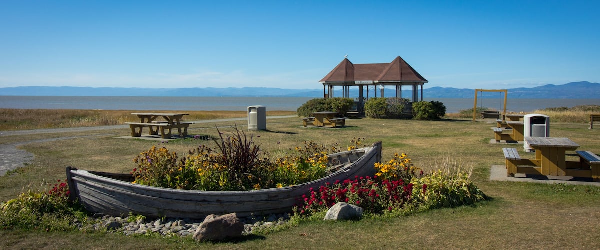 A gazebo, picnic tables, a boat, and wildflowers by a lake with mountains in the background.