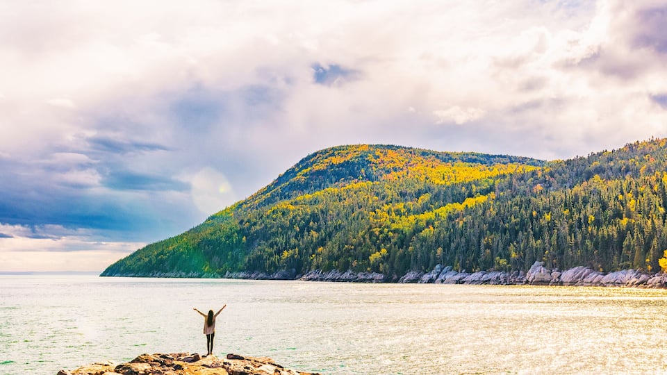 A person standing on a rocky outcrop with arms raised, overlooking a calm lake with a forested mountain in the background.