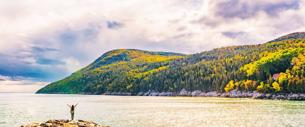 A person standing on a rocky outcrop with arms raised, overlooking a calm lake with a forested mountain in the background.