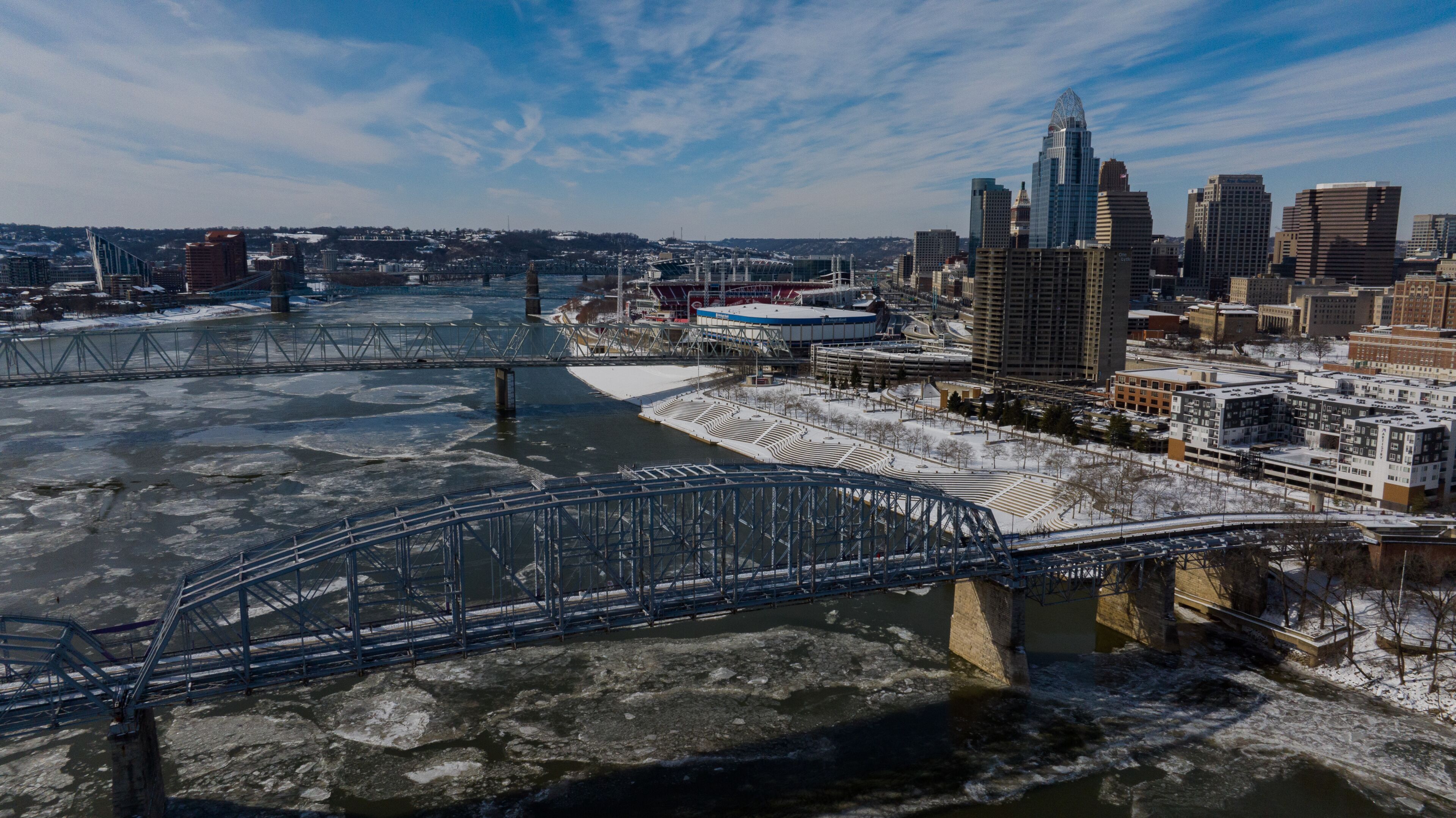 Aerial view of the ice-laden Ohio River flowing beneath bridges towards the Cincinnati skyline and Paycor Stadium under a bright winter sky, Cincinnati, Ohio, United States.