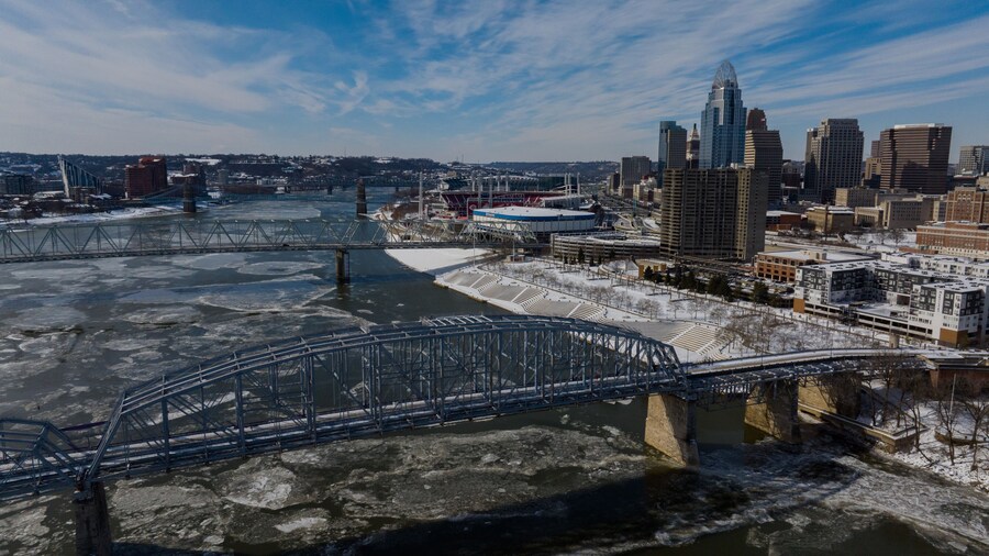 Aerial view of the ice-laden Ohio River flowing beneath bridges towards the Cincinnati skyline and Paycor Stadium under a bright winter sky, Cincinnati, Ohio, United States.