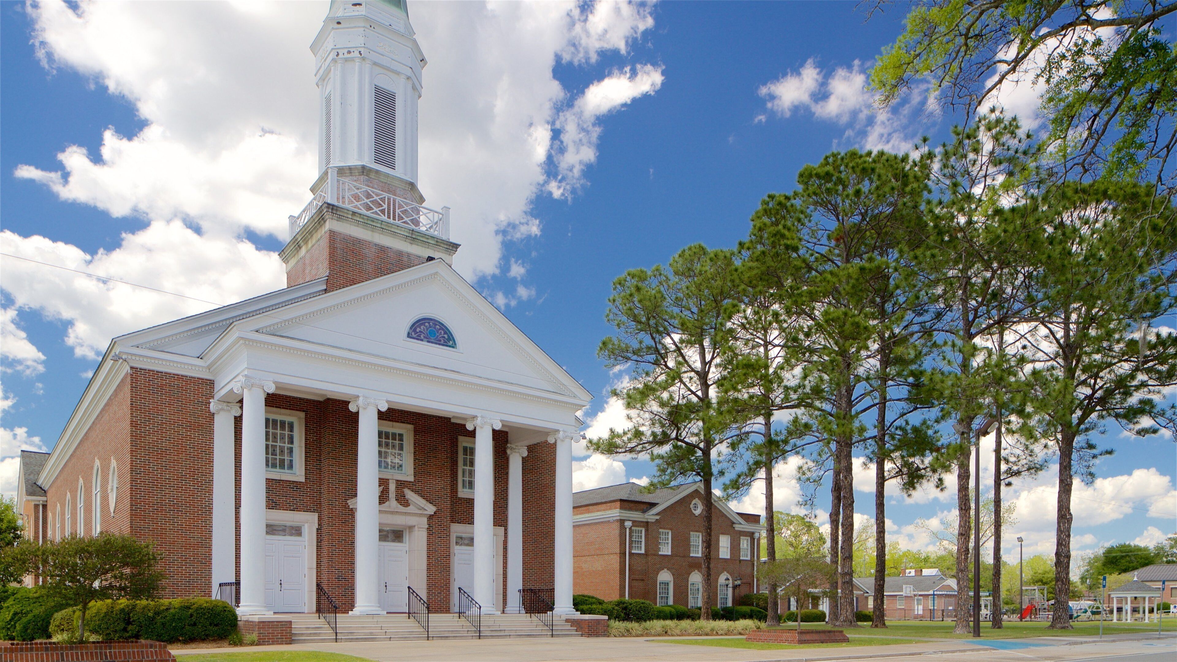 A brick church with a white steeple and columns, surrounded by trees and a clear sky.