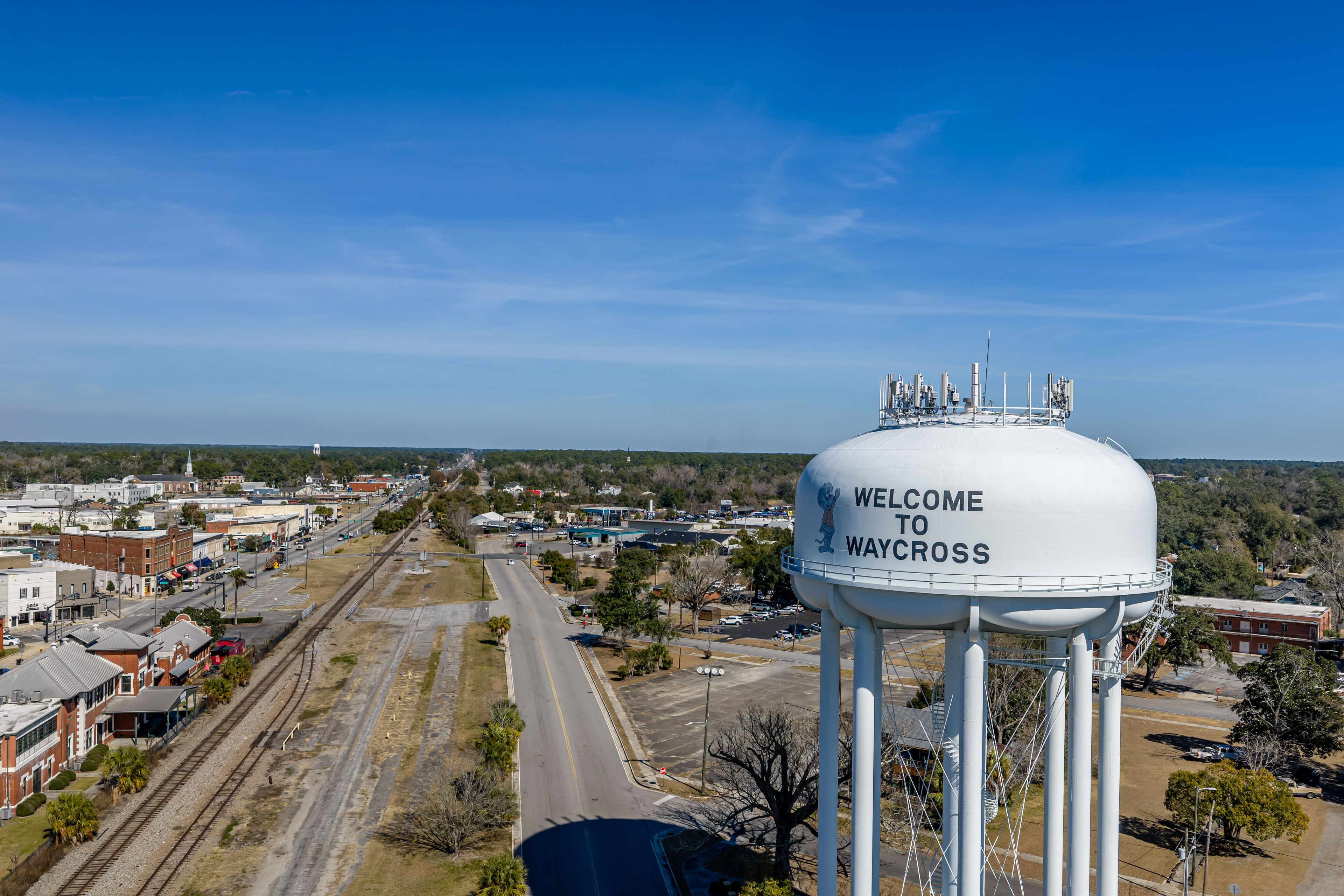 A water tower with "Welcome to Waycross" written on it, overlooking a town with railroad tracks and buildings.