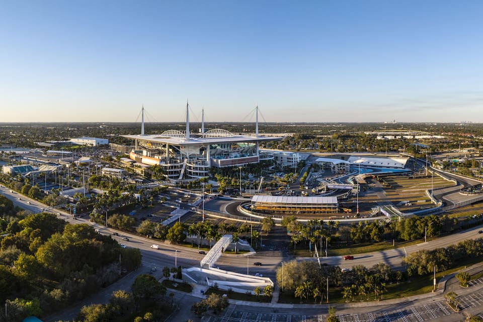 A large stadium with a distinctive roof structure, surrounded by roads and parking areas.