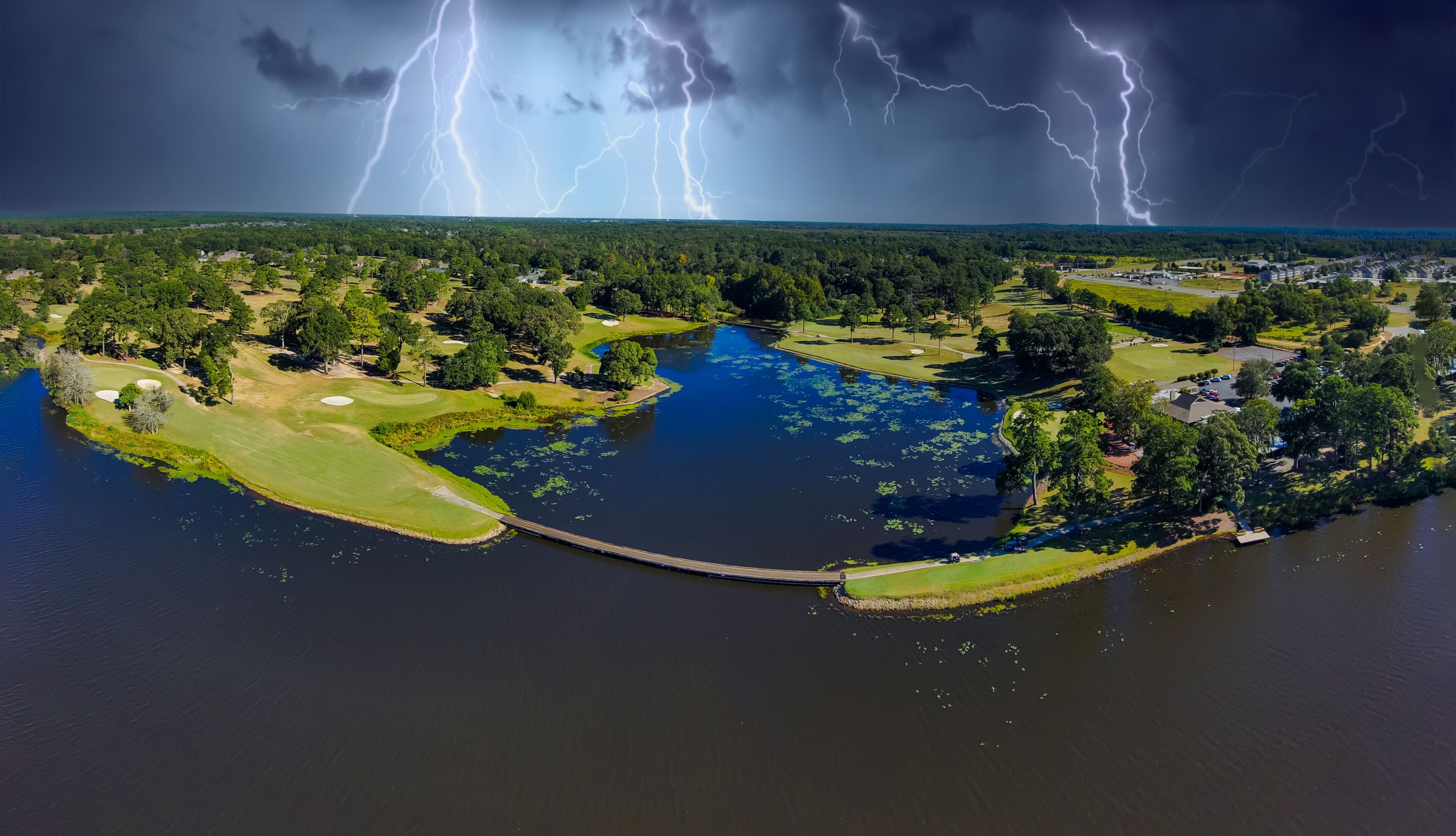 an aerial panoramic shot of a blue waters of Houston Lake surrounded by a golf course and lush green trees, grass and plants with powerful storm clouds and lightning in Perry Georgia USA