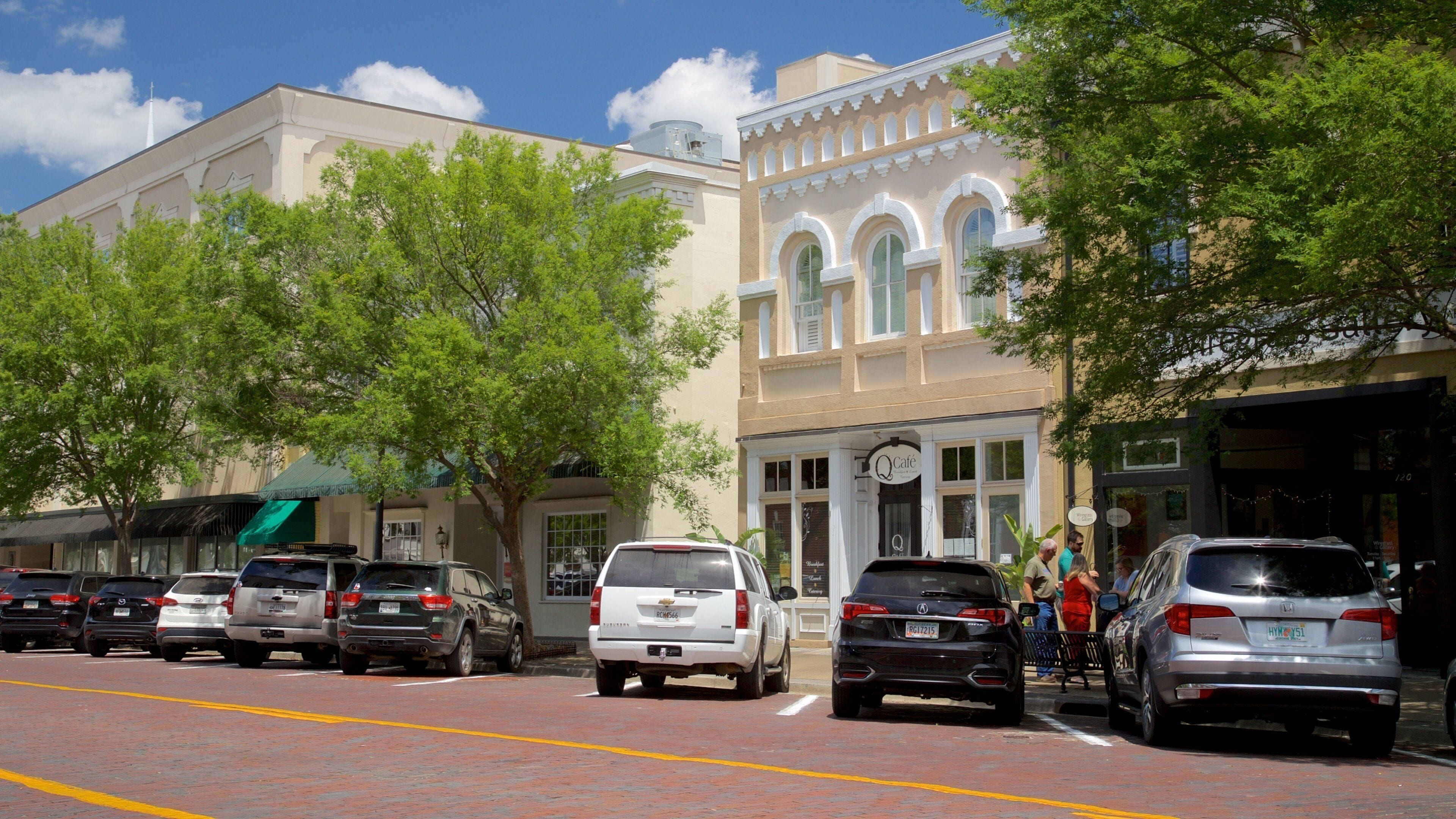 A street lined with parked cars, trees, and buildings with a café.