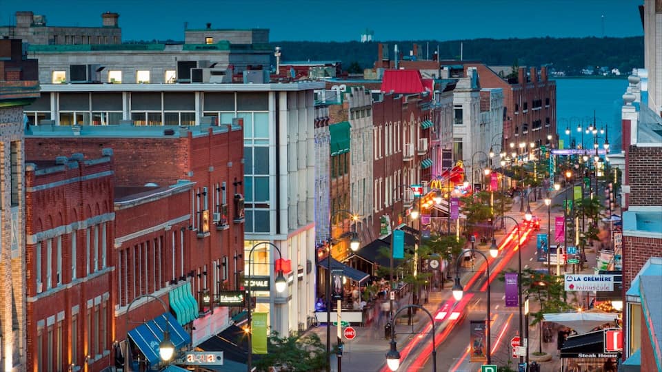 A bustling urban street at dusk with historic buildings, illuminated signs, and a river in the background.
