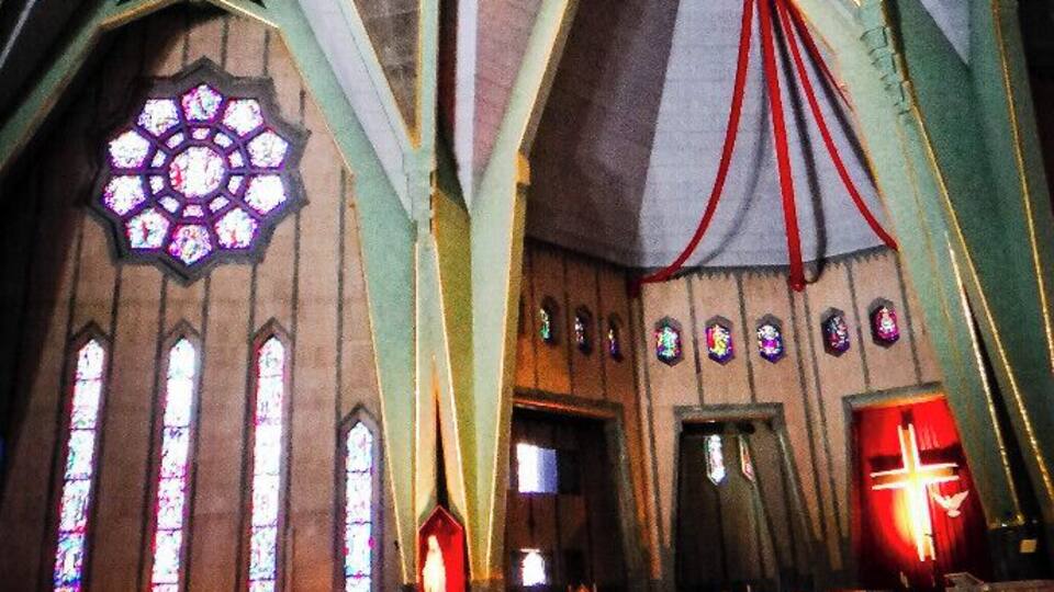 A church interior with a large stained glass window, geometric ceiling design, and a red cross.