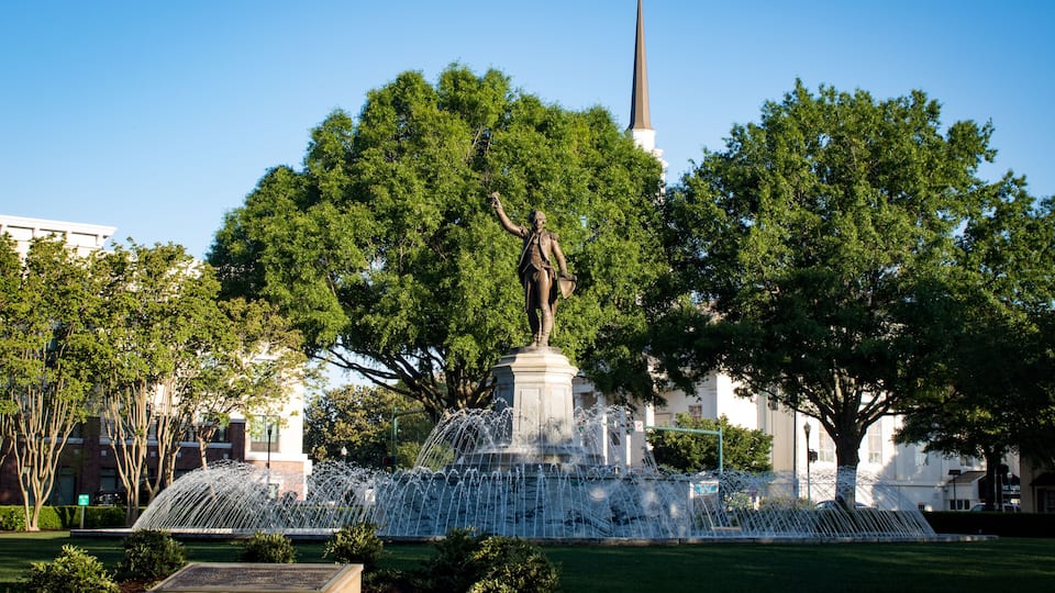 A statue in a park with a fountain, surrounded by trees and a church steeple in the background.