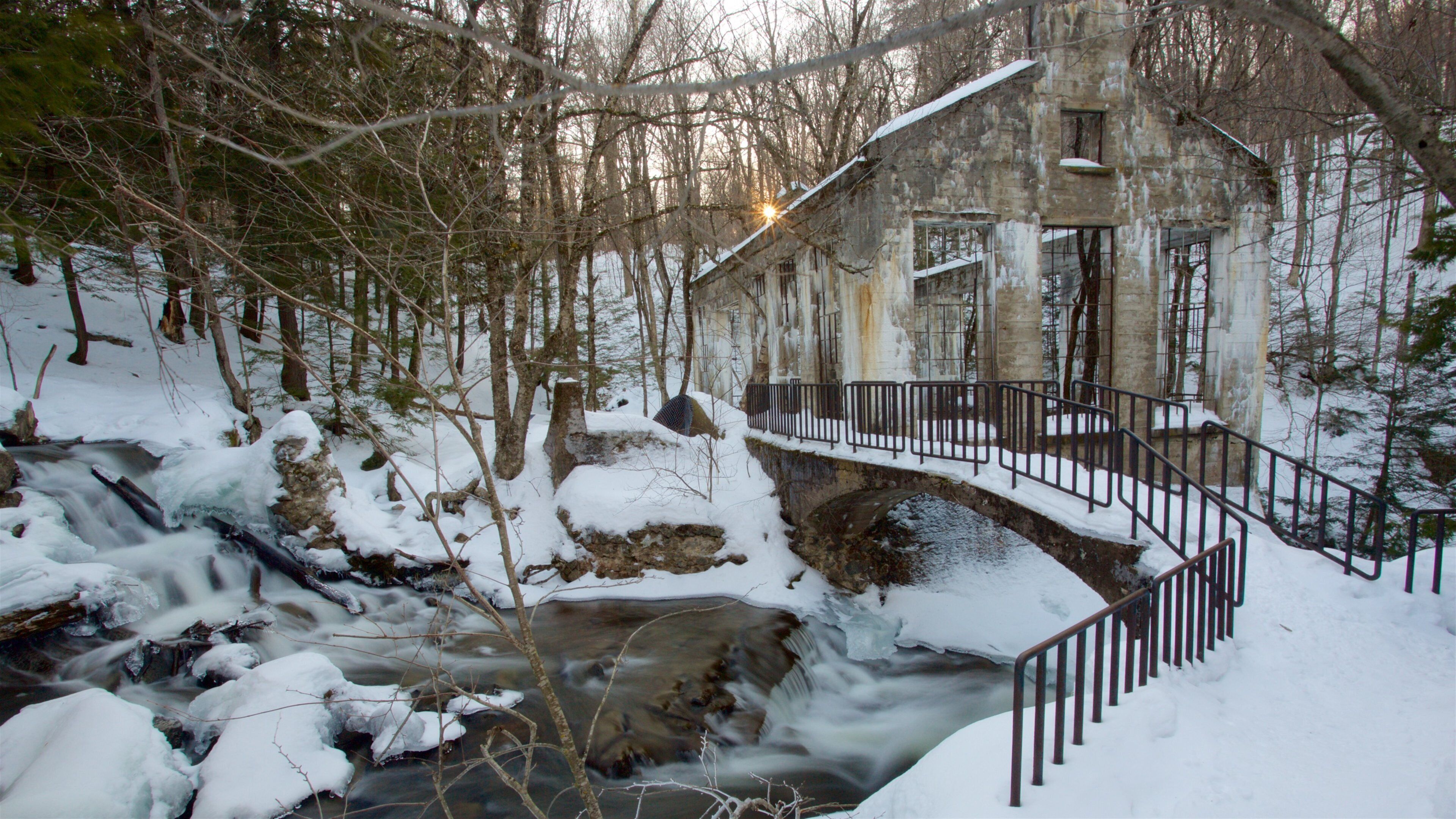 A snow-covered stone bridge over a flowing stream, surrounded by trees and a historic building.