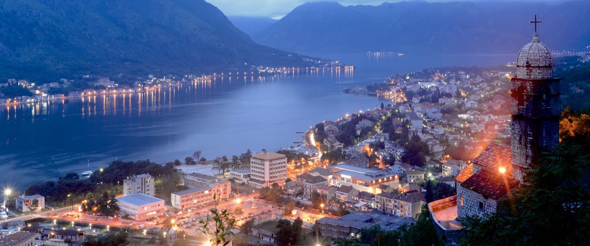 A coastal town with a prominent church tower, a calm lake, and mountains in the background.