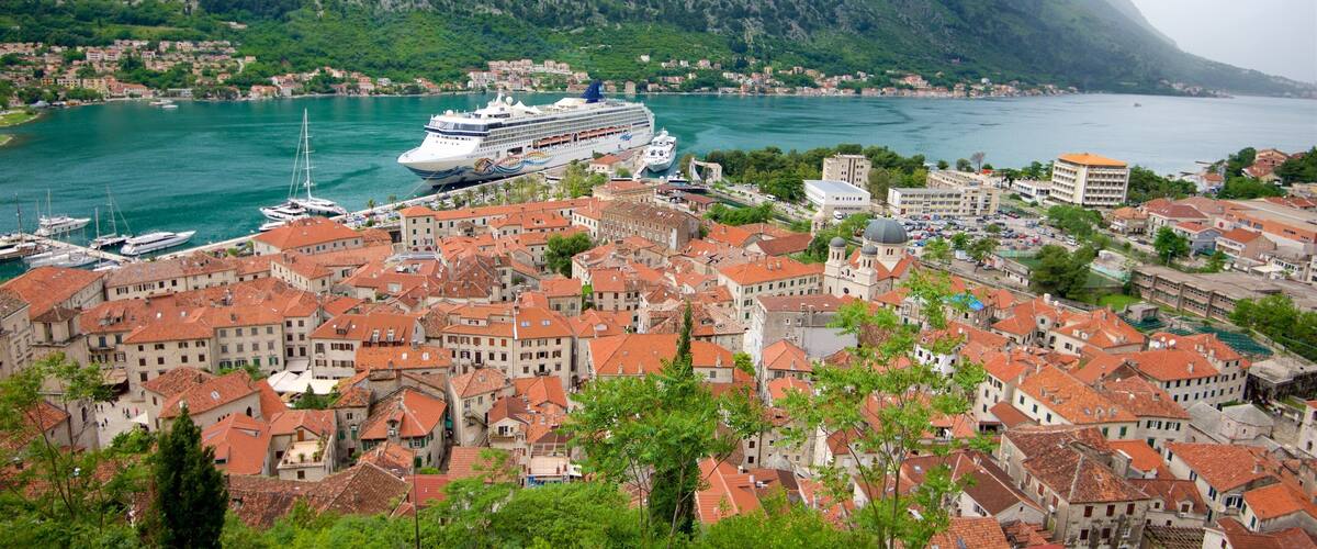 A coastal town with red-roofed buildings, a cruise ship docked at the harbor, and mountains in the background.