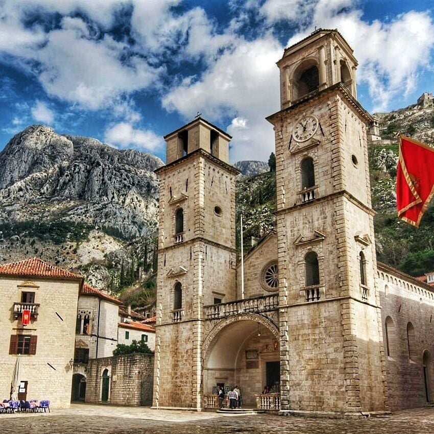 Un edificio de piedra histórico con una torre de reloj, una entrada arqueada y montañas al fondo.
