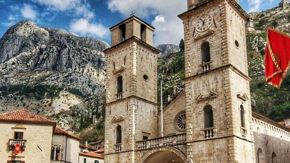 A historic stone building with a clock tower, an arched entrance, and a mountainous backdrop.
