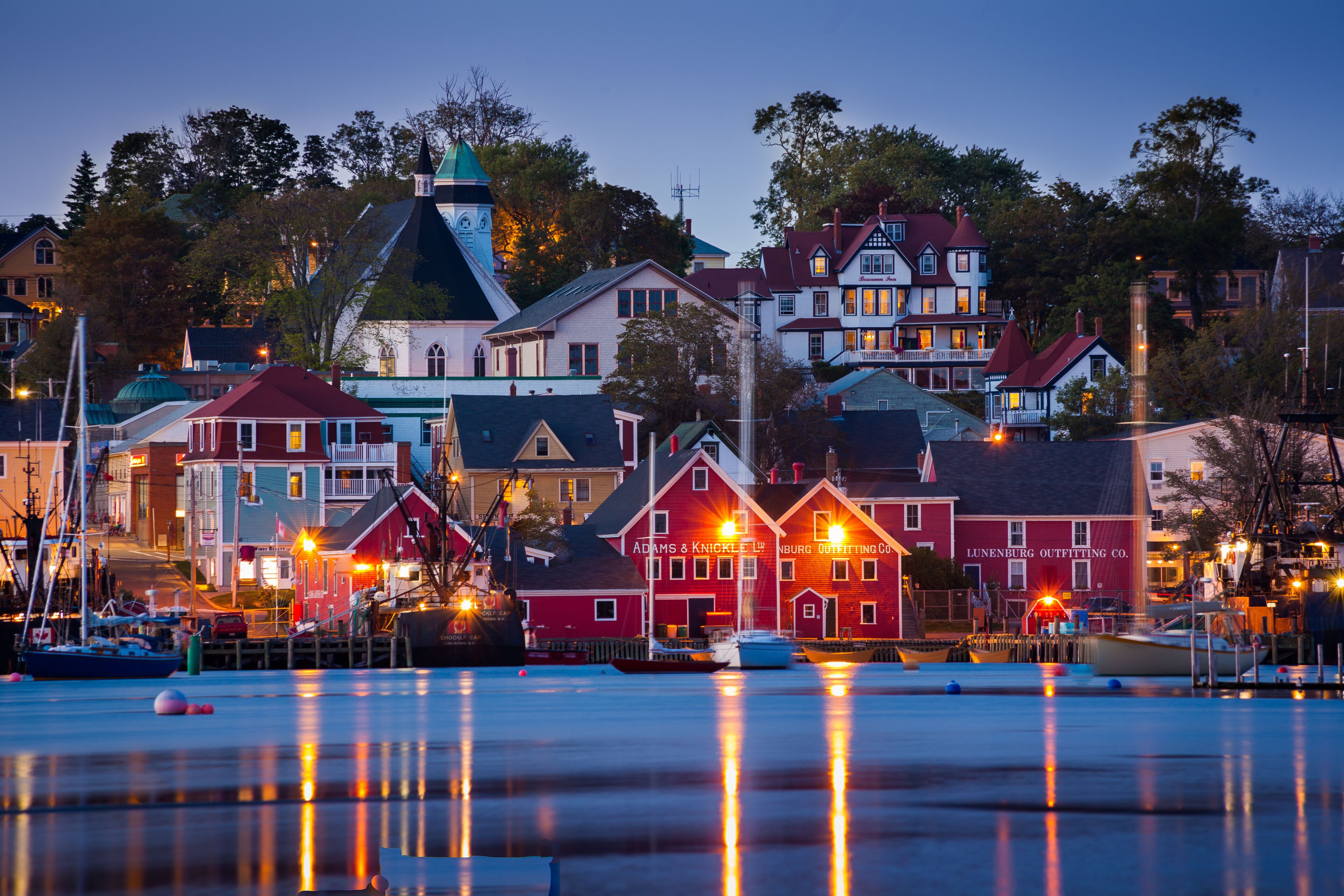 A waterfront town with historic buildings, including a prominent red building with 'Lunenburg Outfitting Co.' signage, and a church with a green roof.