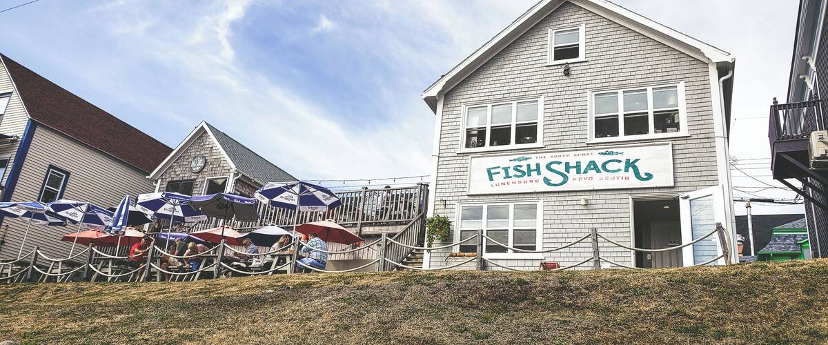 A white building with a sign that reads "Fish Shack" in front of a wooden deck with umbrellas.