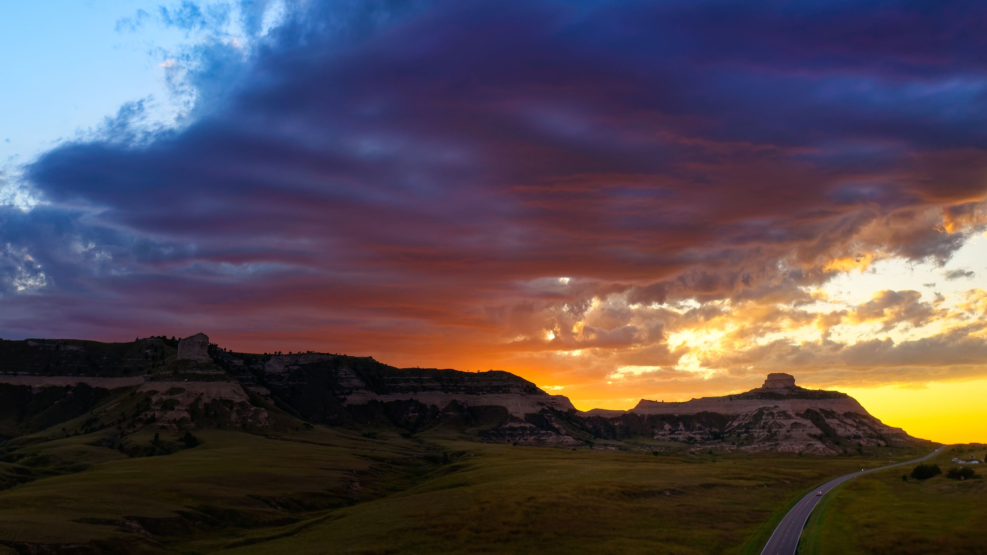 A sunset over rolling hills with a road curving through the landscape.