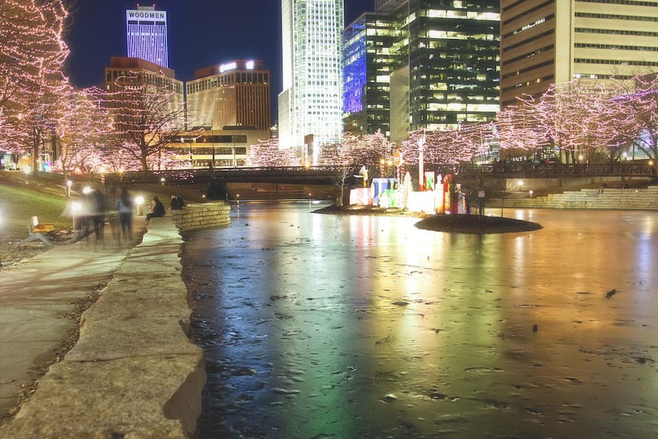 Un paisaje urbano nocturno con edificios iluminados, un río que refleja las luces y árboles de cerezo a lo largo de la orilla.