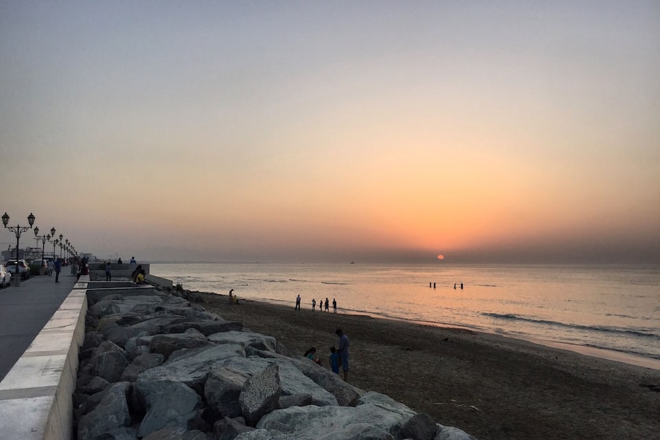 Praia ao pôr do sol, mar calmo, costão rochoso e algumas pessoas caminhando pelo calçadão.