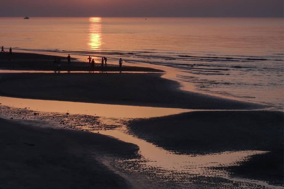 A sunset over a beach with calm waters and people walking along the shore.
