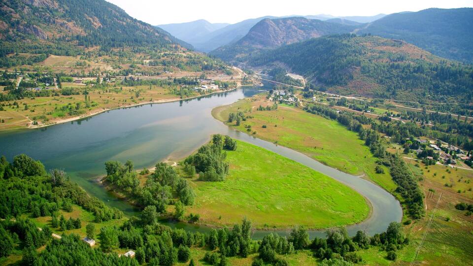A river winding through a valley with a bridge, surrounded by green fields and forested hills.