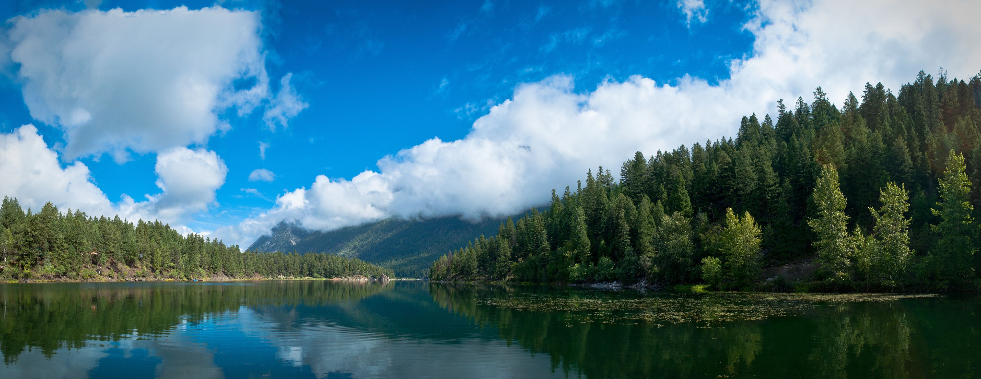 A calm lake surrounded by dense forest and mountains under a partly cloudy sky.