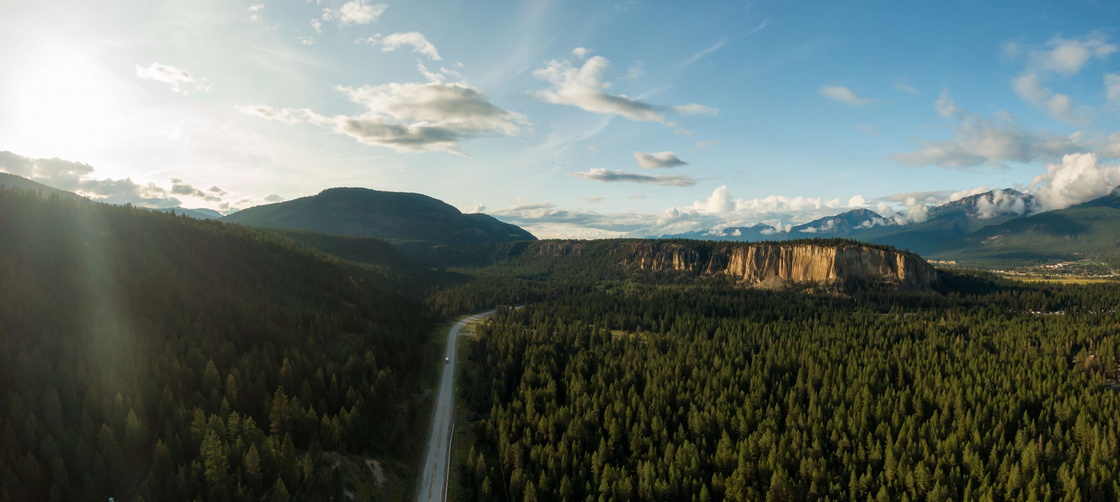 A forested valley with a winding road, mountains in the background, and a clear sky.