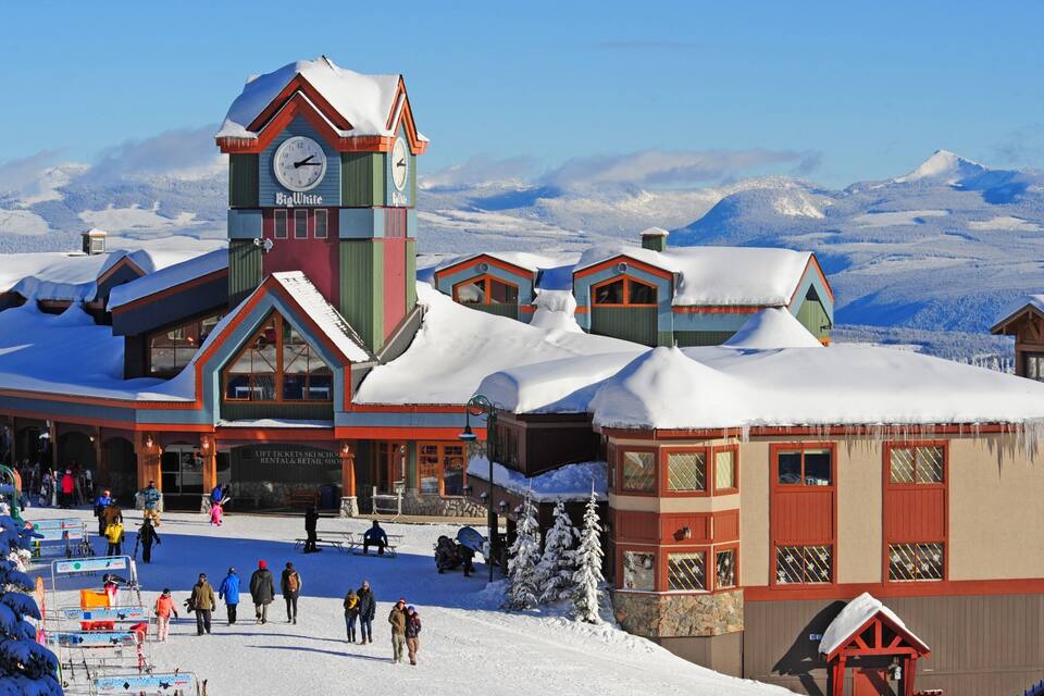 Estación de esquí cubierta de nieve con una imponente torre del reloj, tiendas de alquiler de esquíes y un paisaje montañoso.