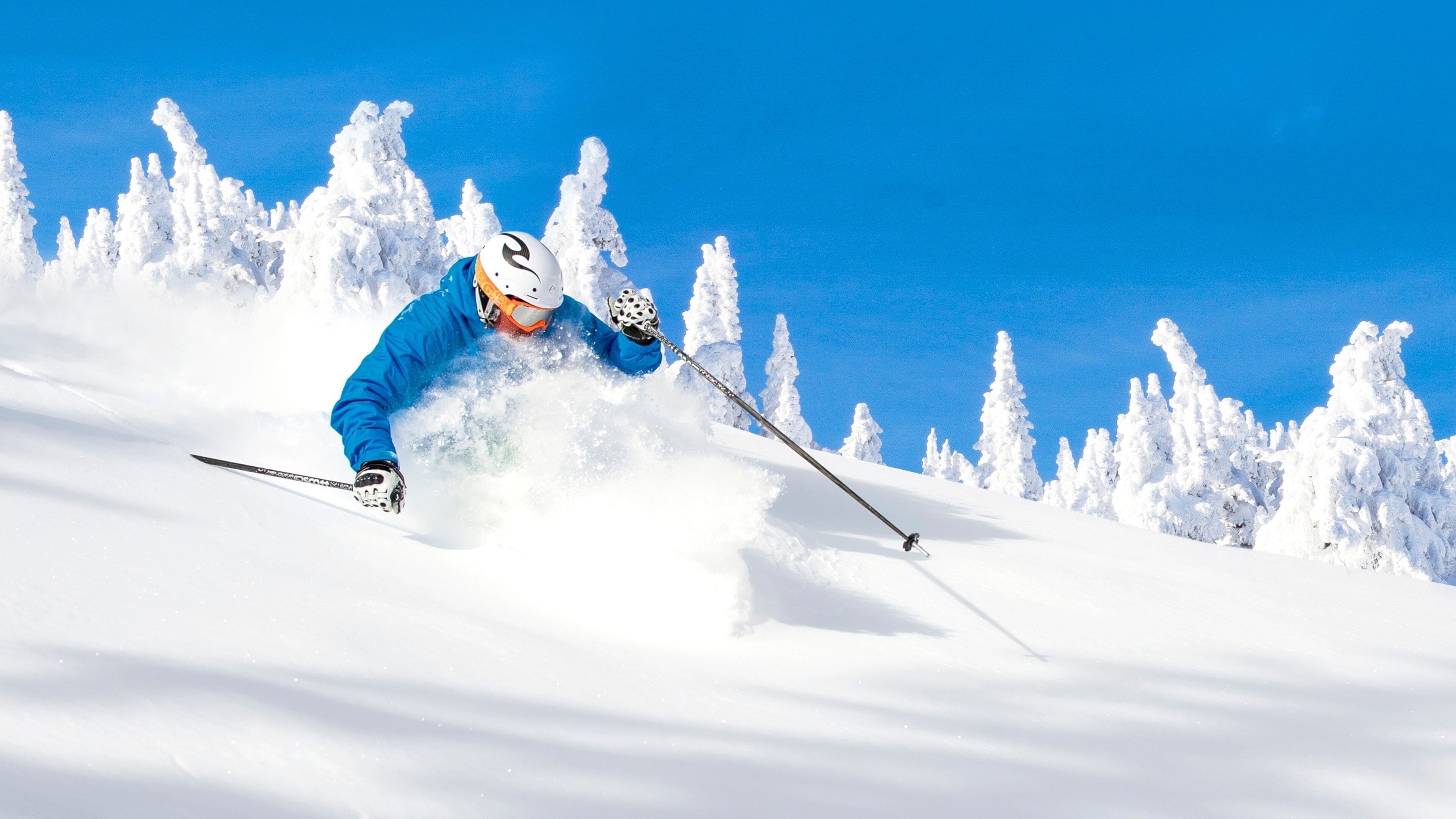 A skier in blue gear carving through deep snow with a clear blue sky in the background.