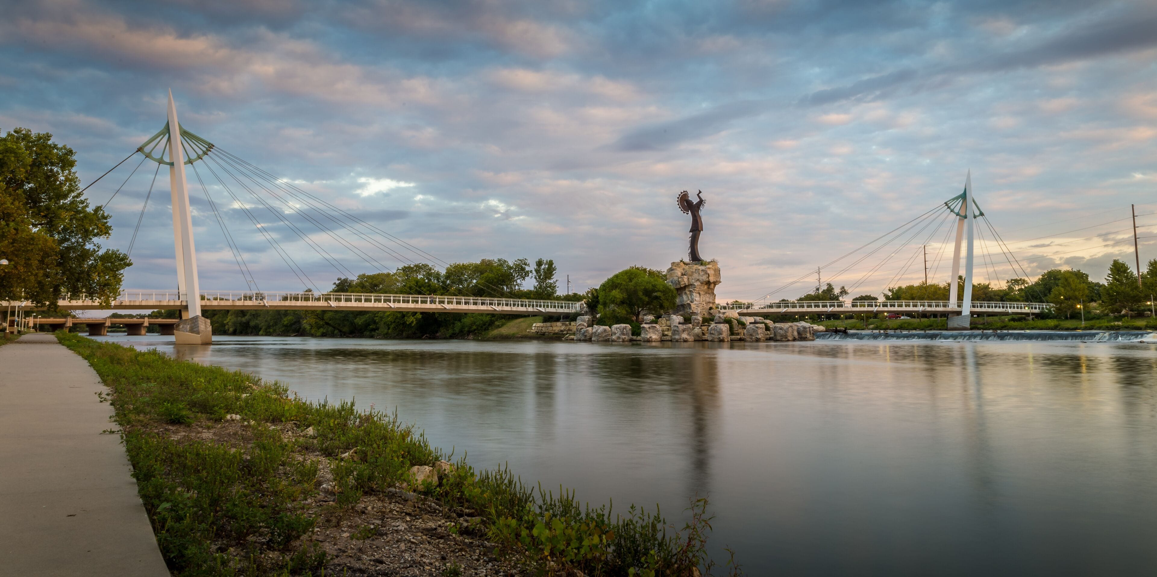 A suspension bridge over a calm river, with a statue on a rock in the center.